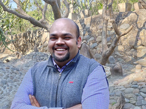 Image of a smiling Indian man, wearing a grey vest over a blue button down shirt, with arms crossed, posing for a picture. He has helped Timberdog, a dog product company, with a carbon sequestration study, to achieve carbon negativity