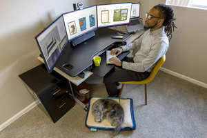 Top-down view of man working at his desk, looking at multiple monitors, typing, while his dog rests on her RuffRest by his feet, dog beds for large dogs, dog bed for small dogs