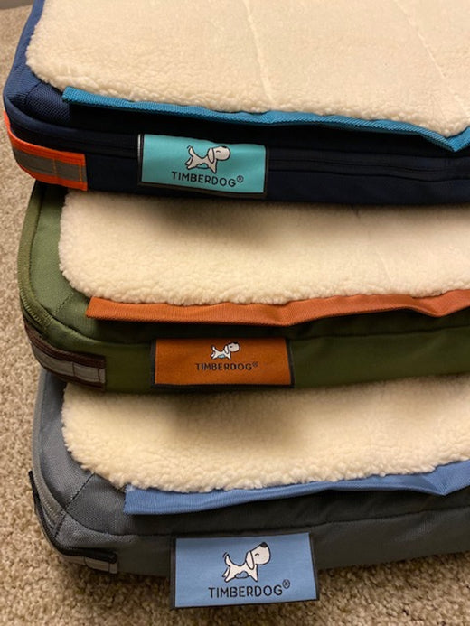 Three dog beds stacked one atop the other in blue, green, and gray colors, with the Timberdog logos or labels clearly visible. Photo taken indoors with dog beds laid out on beige carpet